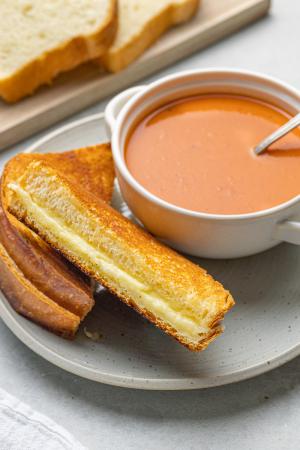A sliced grilled cheese and a bowl of tomato soup sit on a plate in front of a cutting board with more slices of bread.