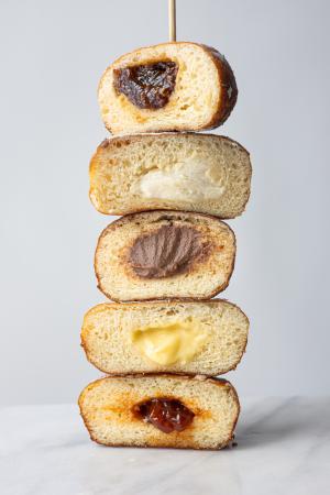 A side view of 5 different flavors of paczki, cut in half to expose the fillings and stacked on top of one another. 