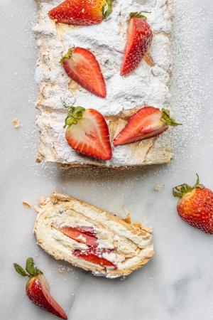 An overhead shot of Eton Mess Roulade dusted with powdered sugar and topped with fresh strawberries. A slice lays on its side to reveal the layers inside.