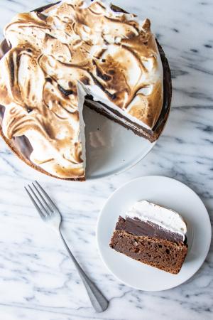 An overhead view of the Mississippi Mud Pie on a white cake stand, featuring a toasted meringue top. Next to it a slice on its side is visible, displaying the brownie, chocolate ganache, and meringue layers. The slice sits on a white plate next to a silver fork, all on a marble counter. 