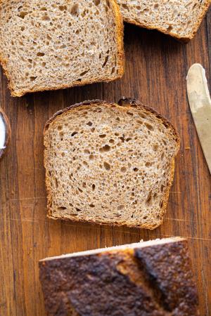 A loaf of naturally leavened bread with 3 slices cut out, laying flat on a wooden surface with a gold knife peeking into frame on the right