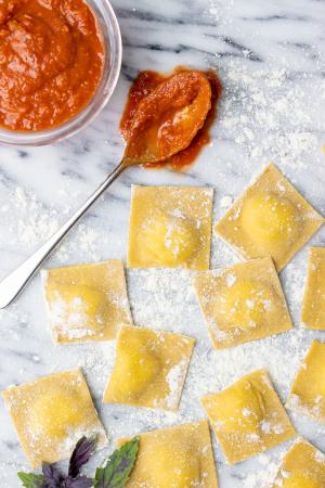 Fresh raviolis sit on a floured marble surface next to a bowl of simple marinara sauce