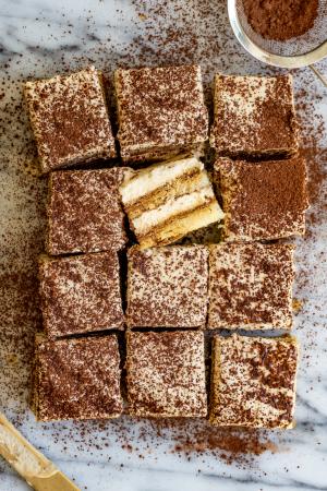 An overhead shot of a tiramisu cut into 12 squares with one square on its side to reveal its layers. A gold knife sits below it and a small mesh sifter filled with cocoa powder sits above it.