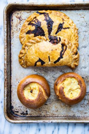 2 pasties sit above 2 knish on a baking sheet atop a marble counter. 