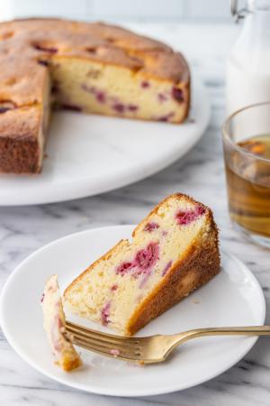 A slice of raspberry ricotta coffee cake next to a cup of tea in front of the rest of the coffee cake on a white cake stand.