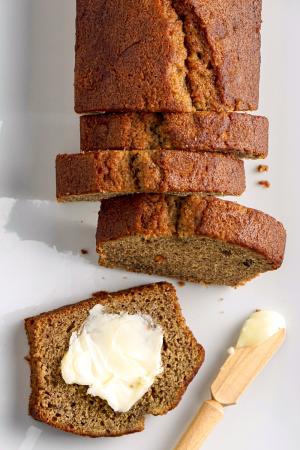 An overheard photo of a loaf of banana bread partially sliced on a white surface. The end slice is laying flat with a smear of butter on it and a small butter spreader next to it.