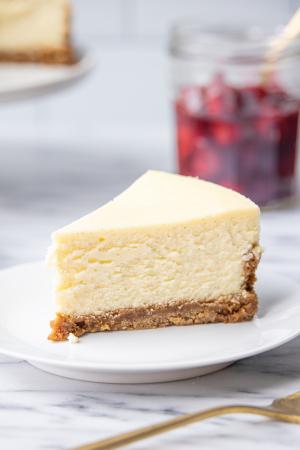 A slice of classic cheesecake on a white plate with a jar of cherries and the rest of the cheesecake on a cake stand visible in the background 
